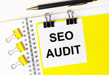 On a light desktop, a pen, yellow paper clips and a white card with the text SEO AUDIT on a yellow background