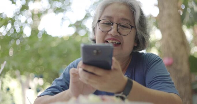 An elderly Asian woman is happily using a smartphone selfie camera to take a picture of food in the garden of her tiny house. Internet communication technology to connect with family