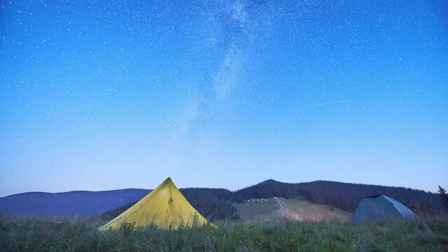 Yellow tent standing against starry sky in mountains. Illuminated tent on hills. Bright light of camping on background of night silhouettes of mountains. Time lapse