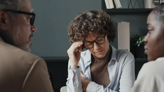 Loose close up of three colleagues at office table, Caucasian senior man and young black woman having serious talk while Caucasian spectacled woman sitting in front of them looking concerned
