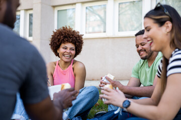 Diverse group of college students. Happy casually dressed multiethnic young friends sitting on the campus grass having a snack while having a conversation.