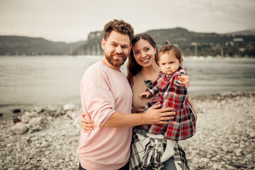 Young happy family spending their vacation on Italian Garda lake coast, bearded daddy, smiling mom, their little toddler girl in red checkered dress pointing with finger