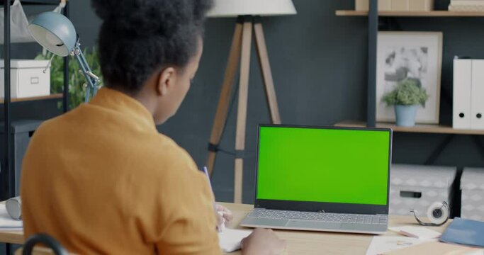 Back view of female employee working with laptop with chroma key green screen writing notes sitting at desk in office. Technology and business concept.