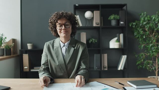 Medium shot of female HR finishing looking through CVs, sitting in light office beside window, then looking at camera, broadly smiling