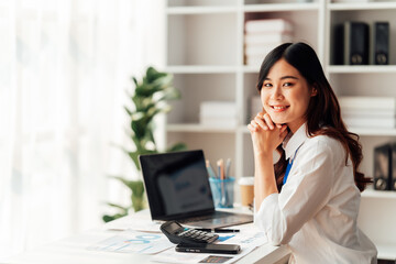 Sharing good business news. Attractive young businesswoman talking on the mobile phone and smiling while sitting at her working place in office and looking at laptop. - obrazy, fototapety, plakaty