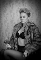 blonde model with pantyhose, glass of red wine ,black bra and fur coat posing provocatively on chair. portrait of sensual blonde, studio shot. Sensual female in black lingerie posing in vintage scene