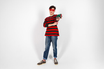A young guy in a red and black clothes and heart shaped glasses posing with a small gift box or a present. Isolated on white background.