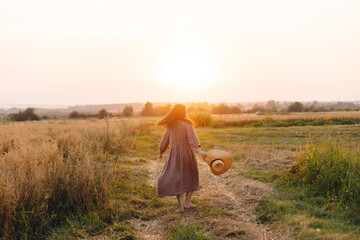 Stylish woman with straw hat dancing at oat field in sunset light. Atmospheric happy moment. Young...