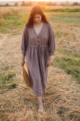 Stylish woman with straw hat walking at oat field in sunset light. Atmospheric tranquil moment....