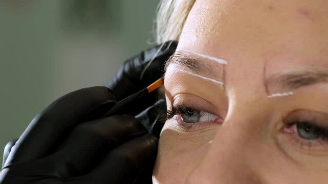Hand of specialist in glove draws white lines near eyebrow of client marking areas on blurred background. Process of brow lamination closeup