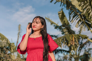 While looking at the sky,smiling, a pretty lady wearing a long red dress is on a phone call with someone . Clear sky and trees in the background