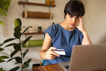 Stressful latin woman with the short hair using credit card at home. She is using laptop and wearing blue dress.