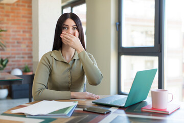 pretty young woman covering mouth with a hand and shocked or surprised expression. laptop and desk concept