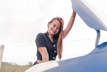 A young adult woman, looking hopeful, and smiling while being stranded on the side of the road and checking the engine in the hood of her blue sedan car