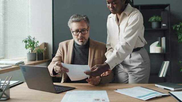 Medium shot of Caucasian senior businessman sitting in office beside window, typing on laptop while his black female colleague bringing him pile of CVs. Then they starting looking through them
