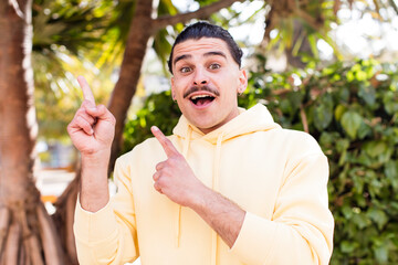 young cool man feeling joyful and surprised, smiling with a shocked expression and pointing to the side