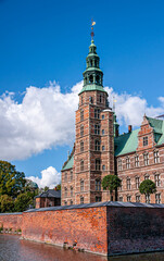 Obraz premium Copenhagen, Denmark - September 13, 2010: Closeup of Rosenborg Slot west red stone facade with tower and green steeple under blue cloudscape. Moat and rampart up front. Green foliage
