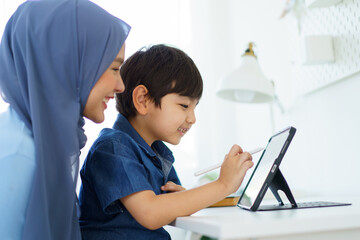 Happy cheerful Asian muslim woman woman teaching her little son a drawing on digital tablet.