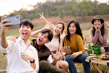 Group of happy Asian young men and women enjoy camping and party together, friends sitting at the park beside lake and enjoy grill a barbecue - stake and playing guitar together.