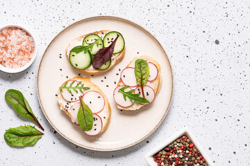 Bright juicy spring and summer snack. Set of bruschetta with cream cheese, cucumbers, small radish and fresh salad leaves on light stone background. Healthy homemade food ideas.