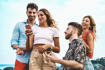 Laughing at Social Media by the Sea - Four young Caucasian friends, two men and two women, near the sea with rocks in the background, laugh while browsing social media on a smartphone.
