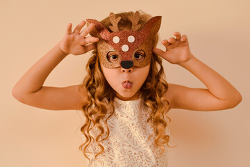 Little cute girl in a carnival masquerade deer mask made of shiny glitter foamiran on a beige...