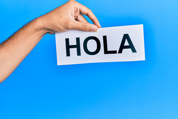 Hand of caucasian man holding paper with hola word over isolated blue background © Krakenimages.com