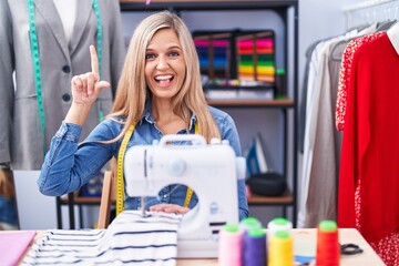 Blonde woman dressmaker designer using sew machine smiling amazed and surprised and pointing up...