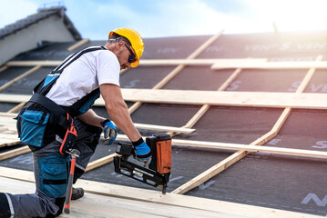 The carpenter nails a timber board using an electric nailer while working on a roof. - obrazy, fototapety, plakaty