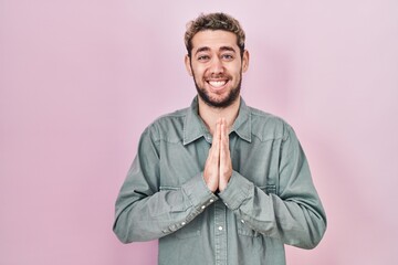 Hispanic man with beard standing over pink background praying with hands together asking for forgiveness smiling confident.