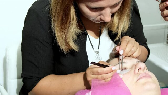 Focused beautician does eyelash extension for young woman using patches and tweezers. Female client with closed eyes enjoys beauty procedure closeup