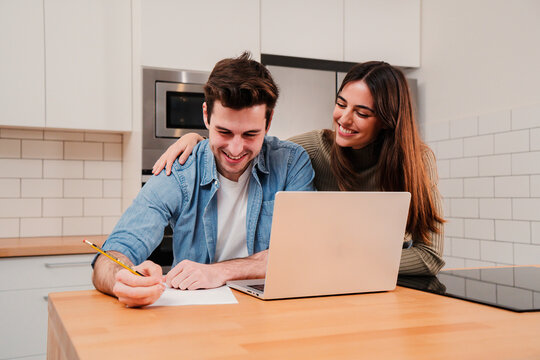Young couple browsing on internet using a laptop searching a mortgage and calculating their budget sitting at home. Handsome man writing the bank accounts on notebook while his wife supports him. High