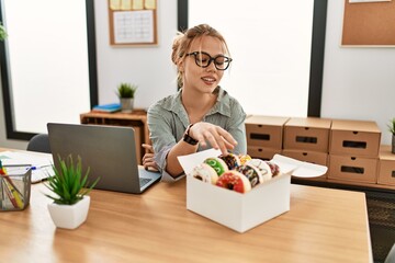 Young caucasian woman business worker using laptop holding doughnut at office