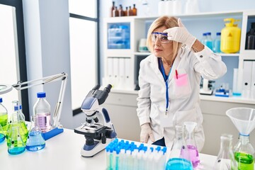 Middle age blonde woman working at scientist laboratory pointing unhappy to pimple on forehead, ugly infection of blackhead. acne and skin problem