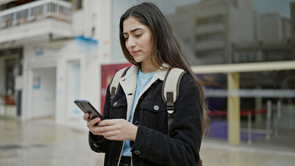 Young beautiful hispanic woman student using smartphone with relaxed expression at street