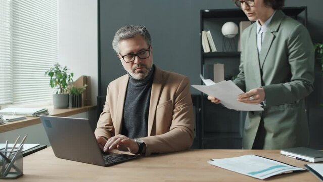 Midsection shot of senior businessman typing on laptop while his female colleague bringing him three CVs. He then starting looking through them, choosing one discuss