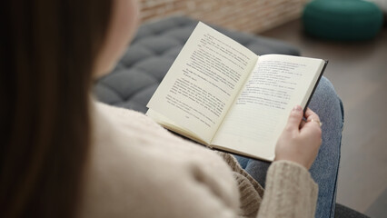 Fototapeta premium Young blonde woman reading book sitting on sofa at home