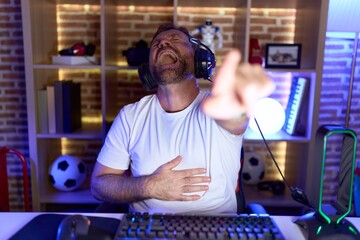 Middle age man with beard playing video games wearing headphones laughing at you, pointing finger to the camera with hand over body, shame expression