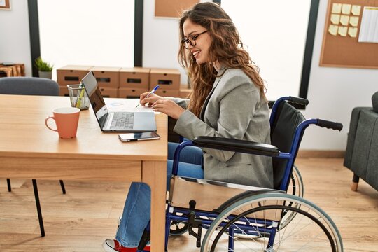 Young beautiful hispanic woman business worker writing on document sitting on wheelchair at office