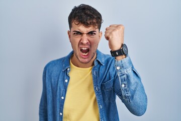 Young hispanic man standing over blue background angry and mad raising fist frustrated and furious...