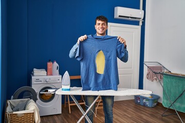 Young hispanic man ironing holding burned iron shirt at laundry room winking looking at the camera...