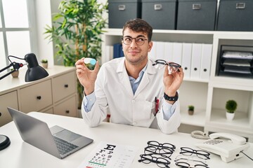 Young hispanic oculist holding glasses and contact lenses smiling looking to the side and staring away thinking.