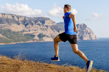male runner running mountain trail along ocean shore, sports clothes blue shirt and black shorts
