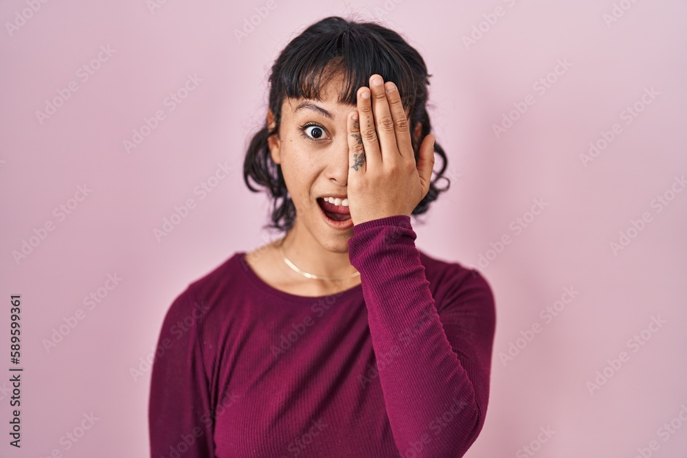 Canvas Prints Young beautiful woman standing over pink background covering one eye with hand, confident smile on face and surprise emotion.