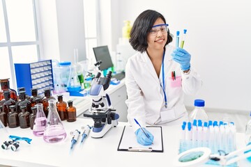 Young latin woman wearing scientist uniform holding test tube writing on clipboard at laboratory