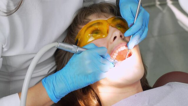 Female dentist and assistant removing dental calculus from teeth. Visit is in proffessional dental clinic. Woman sits on dental chair. Drilling and treatment of tooth, filling.