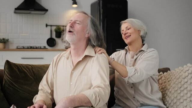 Health, body muscles stiff problem of senior couple, caucasians mature, adult retired husband, wife pain with back pain ache holding massaging rubbing shoulder hurt or sore, painful sitting on sofa.