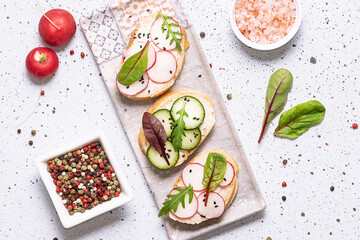 Bright juicy spring and summer snack. Set of bruschetta with cream cheese, cucumbers, small radish and fresh salad leaves on white marble background. Healthy homemade food ideas.