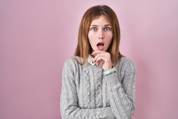 Beautiful woman standing over pink background looking fascinated with disbelief, surprise and amazed expression with hands on chin