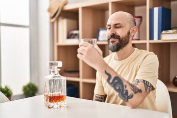 Young bald man drinking glass of whisky sitting on table at home